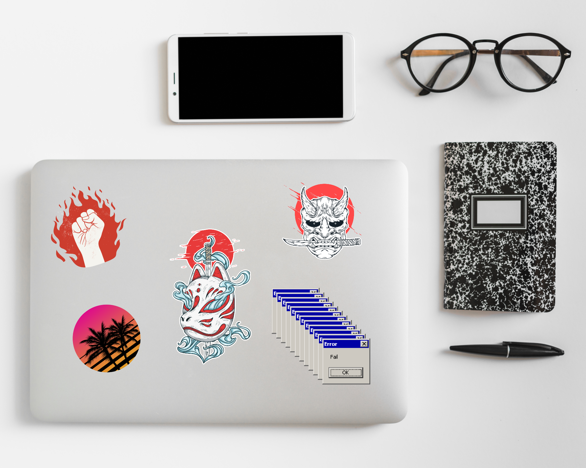 Laptop with stickers, smartphone, eyeglasses, notebook, and pen on a white surface