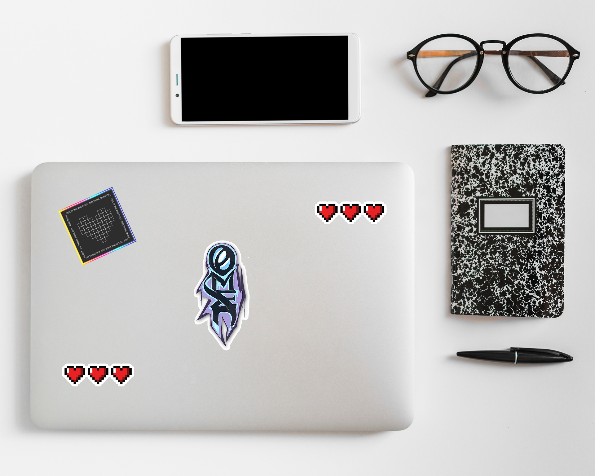 Laptop with stickers, smartphone, eyeglasses, notebook, and pen on a white surface