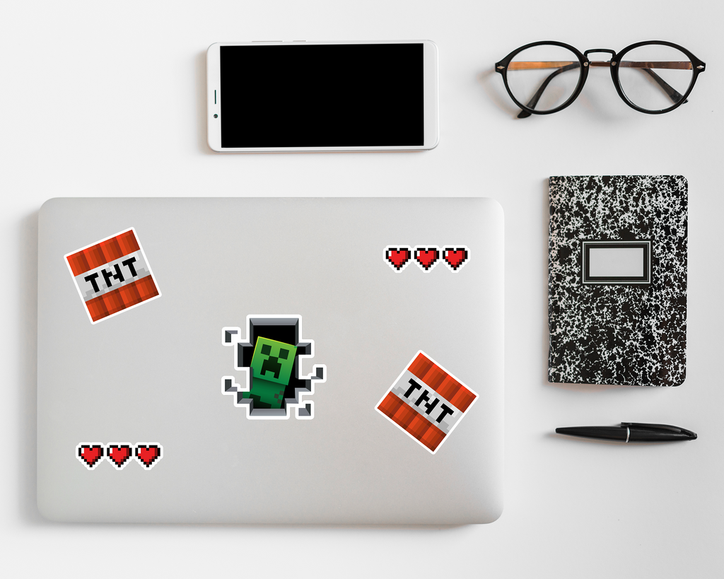 Laptop with Minecraft-themed stickers on a white surface with phone, notebook, and glasses.