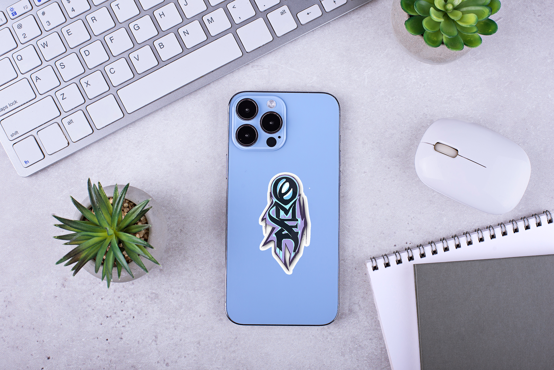 Blue phone case with a omen graffiti sticker on a desk with keyboard, mouse, and plants